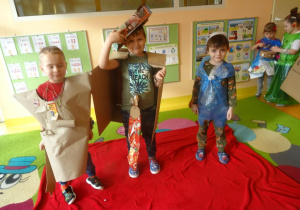 Troje dzieci w ekologicznych strojach stoi na czerwonym dywanie.